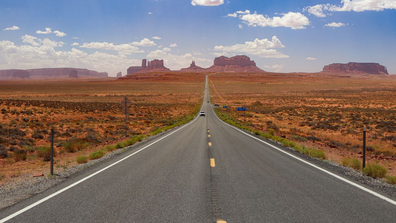 A long, empty highway stretches through Monument Valley under a bright blue sky.